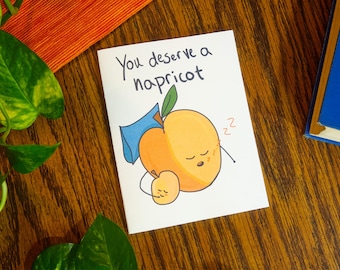 May include: A white greeting card featuring a cartoon illustration of a large orange apricot sleeping with a smaller apricot. The large apricot has a blue pillow behind its head and the text "You deserve a napricot" written in black ink above it.