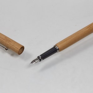 Könnte beinhalten: Ein Holzfüllfederhalter mit abgenommener Kappe auf einer weißen Oberfläche. Der Stift hat einen hellbraunen Holzschaft und eine Kappe, einen schwarzen Griffbereich und eine silberne Feder. Die Kappe befindet sich links mit einem silbernen Clip.