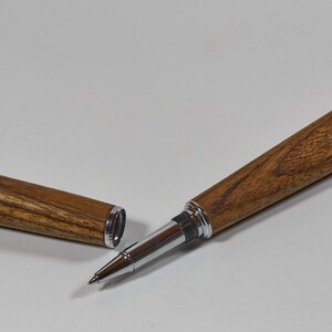 Könnte beinhalten: Ein Holz-Rollerball-Stift mit seiner Kappe daneben. Der Stift hat einen braunen Holzmaserungslauf mit silberfarbenen Metallakzenten. Die Kappe ist ebenfalls aus Holz mit silberfarbenen Akzenten. Der Stift liegt auf einer weißen Oberfläche.