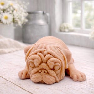 A detailed handmade vegan soap gift, sculpted in the shape of a pug dog lying flat on its stomach in a sleeping position, showing realistic skin folds and fur texture, displayed on a light wooden surface. Handmade in Blackpool