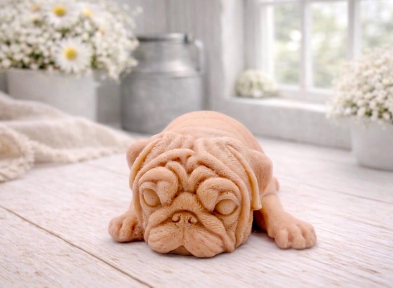 A detailed handmade vegan soap gift, sculpted in the shape of a pug dog lying flat on its stomach in a sleeping position, showing realistic skin folds and fur texture, displayed on a light wooden surface. Handmade in Blackpool