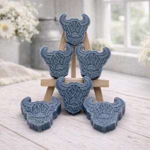 A collection of six handmade vegan soaps each intricately sculpted into the head of a shaggy Highland cow with long horns and a thick, textured coat, arranged in a pyramid on and around a small wooden easel.