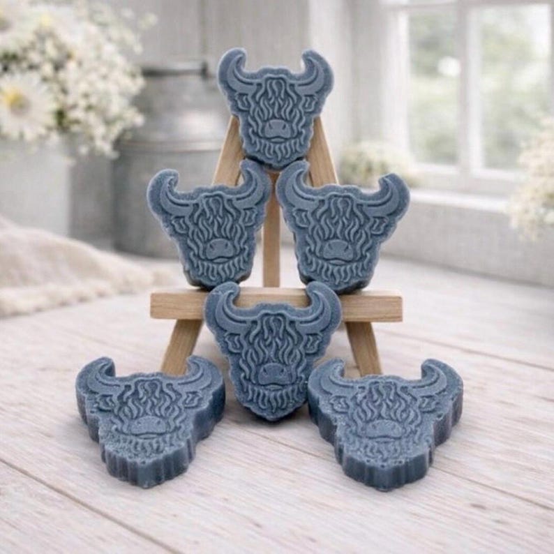 A collection of six handmade vegan soaps each intricately sculpted into the head of a shaggy Highland cow with long horns and a thick, textured coat, arranged in a pyramid on and around a small wooden easel.
