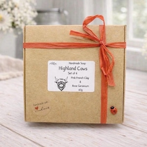 A collection of six handmade vegan soaps each intricately sculpted into the head of a shaggy Highland cow with long horns and a thick, textured coat, arranged in a pyramid on and around a small wooden easel.