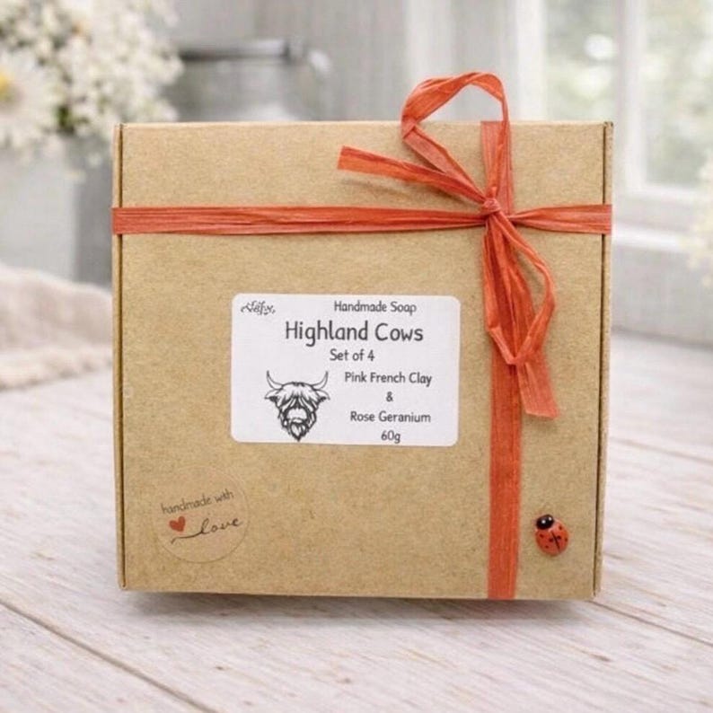 A collection of six handmade vegan soaps each intricately sculpted into the head of a shaggy Highland cow with long horns and a thick, textured coat, arranged in a pyramid on and around a small wooden easel.
