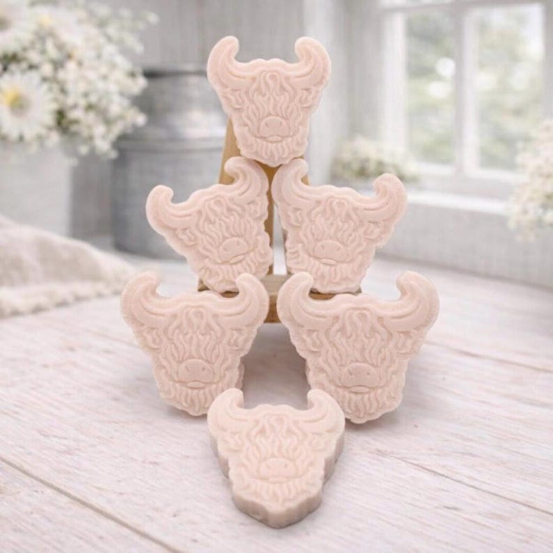 A collection of six handmade vegan soaps each intricately sculpted into the head of a shaggy Highland cow with long horns and a thick, textured coat, arranged in a pyramid on and around a small wooden easel.