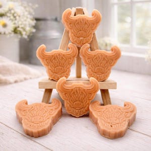 A collection of six handmade vegan soaps each intricately sculpted into the head of a shaggy Highland cow with long horns and a thick, textured coat, arranged in a pyramid on and around a small wooden easel.