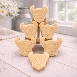 A collection of six handmade vegan soaps each intricately sculpted into the head of a shaggy Highland cow with long horns and a thick, textured coat, arranged in a pyramid on and around a small wooden easel.