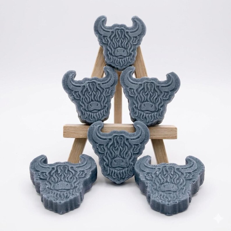 A collection of six handmade vegan soaps each intricately sculpted into the head of a shaggy Highland cow with long horns and a thick, textured coat, arranged in a pyramid on and around a small wooden easel.