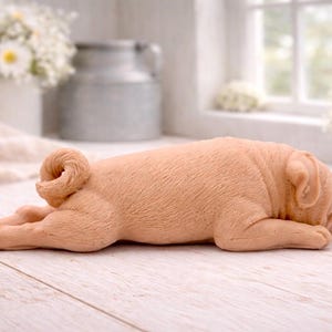 A detailed handmade vegan soap gift, sculpted in the shape of a pug dog lying flat on its stomach in a sleeping position, showing realistic skin folds and fur texture, displayed on a light wooden surface. Handmade in Blackpool