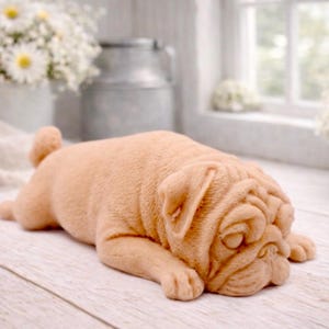 A detailed handmade vegan soap gift, sculpted in the shape of a pug dog lying flat on its stomach in a sleeping position, showing realistic skin folds and fur texture, displayed on a light wooden surface. Handmade in Blackpool