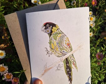 Op de afbeelding: Een wenskaart met een gedetailleerde illustratie van een papegaai die op een tak zit. De papegaai is versierd met bloemmotieven in geel, groen en rood. De kaart wordt vastgehouden met een bruine envelop tegen een achtergrond van madeliefjes.