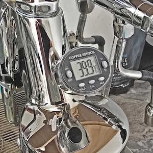 Peut inclure: Gros plan sur une machine à café avec un capteur de température numérique. Le capteur affiche 39,9°F (4,4°C). La machine est en chrome brillant et comporte divers tuyaux et buses. Le capteur comporte des boutons "ON", "OFF", "HOLD", "CAL" et "MAX".