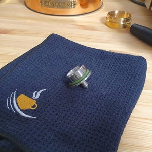 May include: A silver and green metal espresso machine part on a dark blue towel. The towel has a white embroidered coffee cup design.