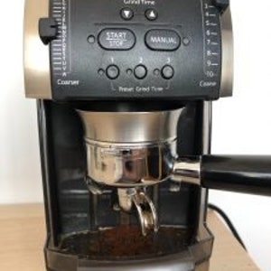 Może przedstawiać: Czarny i srebrny młynek do kawy Vario z cyfrowym wyświetlaczem i przyciskami sterowania. Młynek ma zamontowany portafiltr, gotowy do przyjęcia zmielonej kawy. Widoczne są napisy "START/STOP" i "MANUAL".