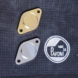 Op de afbeelding: Twee metalen, ruitvormige, espressomachine portafilter blanco's. Eén is goud en de andere is zilver. De blanco's liggen op een donkergrijze stoffen achtergrond. Een wit geborduurd logo met de tekst "Pavoni" en een kroon is zichtbaar op de achtergrond.