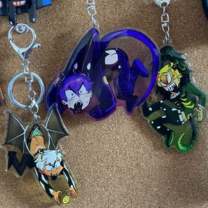 Puede incluir: Varios llaveros acrílicos con diseños de personajes de dibujos animados. Uno muestra una criatura parecida a un murciélago con alas. Otro presenta un personaje de pelo morado con un atuendo oscuro. Un tercer llavero presenta un monstruo verde y amarillo.