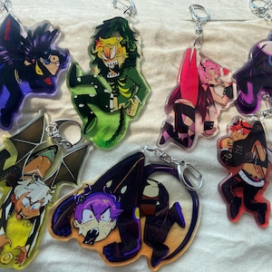 Peut inclure: Une collection de six porte-clés en acrylique représentant des personnages de style anime dans diverses poses. Les motifs incluent des couleurs vives comme le violet, le vert, le rose, le jaune et le noir, chacun fixé à un mousqueton en métal argenté.