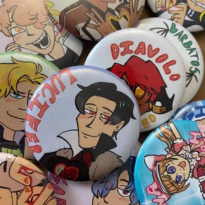 Op de afbeelding: Een verzameling kleurrijke, ronde buttons met illustraties van personages in anime-stijl. De buttons tonen verschillende personages met unieke ontwerpen en tekst, waaronder de namen "Lucifer" en "Diavolo". De buttons zijn in verschillende kleuren, waaronder blauw, rood en wit.