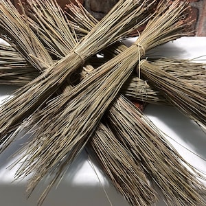 Può includere: Diversi fasci di aghi di pino secchi sono disposti a croce. Ogni fascio è legato con spago. Gli aghi sono un misto di verde chiaro e scuro, con alcune punte marroni. Lo sfondo è un muro di mattoni.