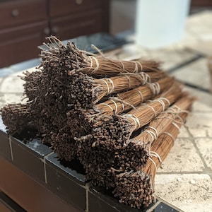 May include: Several bundles of dried pine needles, secured with twine and elastic bands. The needles are a rich brown hue, and the bundles are stacked on a tiled surface. The background is blurred, possibly indicating a kitchen environment.