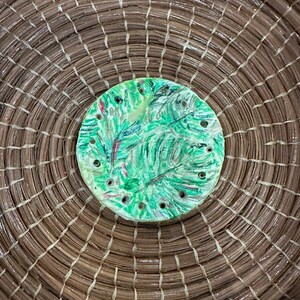 Peut inclure: Un panier rond tissé avec un élément décoratif central circulaire. L'élément est peint en vert avec un motif de feuilles et de petits trous. Le panier est fait de fibres naturelles brunes.