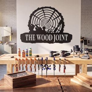 May include: A black metal wall sign featuring a circular tree ring design with woodworking tools and the text THE WOOD JOINT. The sign is mounted on a white wall above a wooden workbench equipped with various clamps, glue bottles, and power tools.