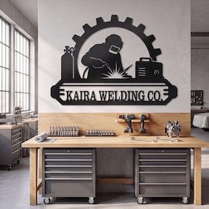 May include: A black metal wall sign featuring a silhouette of a welder wearing a helmet, a gas cylinder, and a welding machine, all framed by a gear shape. Below the central design, a horizontal banner displays the text KAIRA WELDING CO.