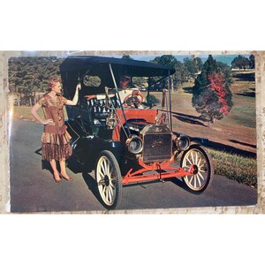May include: Vintage Ford Model T car with a black top, red accents, and cream-colored wheels. A woman in a brown dress stands next to the car on a paved road. The background features trees and a grassy area.