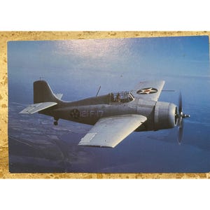 May include: A vintage photograph of a gray military aircraft in flight over a blue ocean. The plane has a propeller, wings, and a tail. The aircraft has the number "121-F-17" printed on the side and a star emblem.