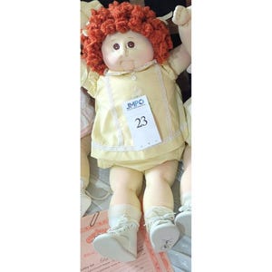 May include: A vintage Cabbage Patch Kid doll with a yellow dress and red curly hair. The doll has a tag that reads "JMPO 23". The doll is wearing white shoes and socks. A classic collectible toy.