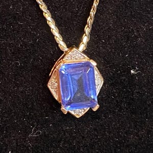 May include: A gold-toned necklace with a rectangular, deep blue gemstone pendant. The pendant is surrounded by a diamond-studded frame. The chain has a braided design. The jewelry is set against a black background.