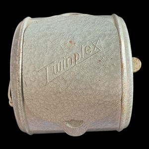 May include: A vintage, cylindrical object with a light gray, textured surface. The word "Twinplex" is printed on the side. It has a small, round, gold-colored knob and a small opening on the bottom. The object appears to be made of metal.