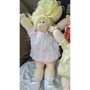 May include: A vintage doll with blonde yarn hair, wearing a pink dress with floral embroidery and white shoes. The doll has a tag attached. The doll's arms are outstretched. Another doll is partially visible on the right.