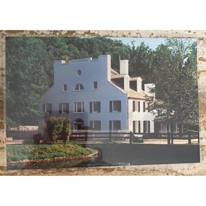 May include: A photograph of a light blue and white historic house with multiple windows and a white fence. The house is surrounded by green trees and a small body of water. The sky is overcast.