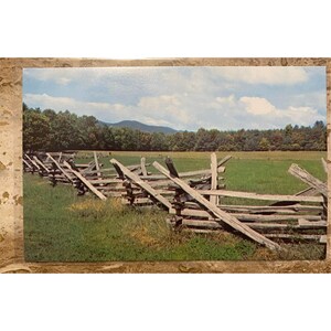 May include: A vintage postcard featuring a split-rail fence in a green field, with a backdrop of trees and a mountain range under a cloudy sky. The fence is made of weathered wood, and the scene evokes a sense of rural tranquility.