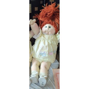 May include: A vintage doll with a round face, green eyes, and bright orange yarn hair. The doll wears a light yellow dress with a baby carriage design and white shoes. The doll is a collectible item.