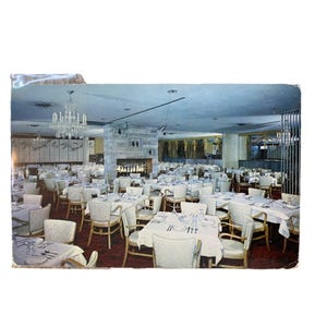 May include: An interior shot of a vintage restaurant or banquet hall. Numerous tables are set with white tablecloths, silverware, and glassware. The room features a fireplace, a chandelier, and decorative curtains, suggesting a formal dining setting.