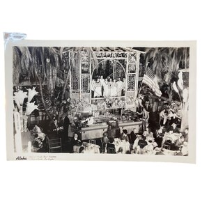 May include: Black and white photograph of a vintage restaurant scene. The word "Aloha" is visible above a stage where people are standing. Palm trees and an American flag are also visible.