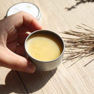 May include: A small, open, silver-colored tin filled with a yellow balm. The tin is held in a hand, with another tin in the background. Dried plants and a wooden surface complete the scene.