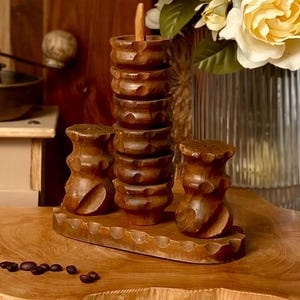 Peut inclure: Un ensemble de salière et poivrière en bois avec un support assorti. L'ensemble comprend deux salières et un grand récipient avec une cuillère en bois, le tout dans une finition marron foncé. Les articles sont posés sur un plateau en bois avec un bord festonné.