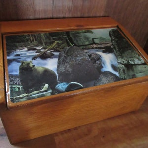 May include: A wooden box with a hinged lid. The lid features a colorful image of a bear in a forest setting with a waterfall. The box is made of natural wood with a light brown finish.