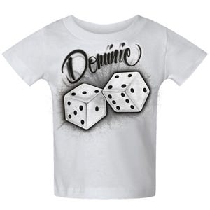 May include: White t-shirt featuring an airbrushed design of two dice and the name "Dominic" in a stylized font. The dice are white with black dots, set against a gray, smoky background. The shirt is a classic crew neck style.