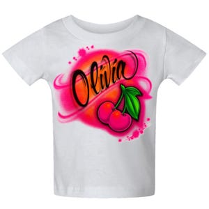 May include: White t-shirt featuring the name "Olivia" in black script, set against a pink and orange airbrushed background. Below the name are two pink cherries with a green leaf.