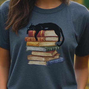 Puede incluir: Camiseta azul marino con un diseño de un gato negro durmiendo sobre una pila de libros coloridos. Los libros tienen lomos rojos, amarillos, verdes y azules. El gato está representado en una pose relajada.