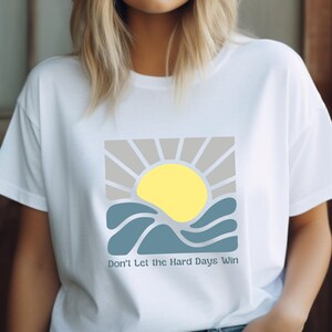 以下が含まれることがあります： 黄色い太陽、灰色の光線、青い波のグラフィックが特徴の白いTシャツ。「Don't Let the Hard Days Win」の文字が下にプリントされています。このシャツは柔らかい素材で作られています。