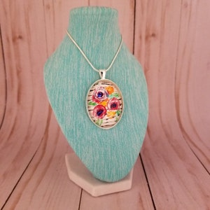 Puede incluir: Un collar de cadena de plata con un colgante redondo que presenta un diseño floral pintado. El colgante tiene un fondo blanco con flores rosas, rojas, amarillas y azules.