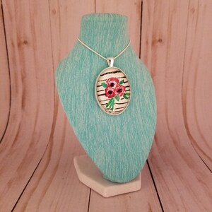 Puede incluir: Un collar de cadena de plata con un colgante redondo con un fondo a rayas blanco y negro con un diseño floral rojo y verde.