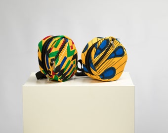 Può includere: Due borse rotonde in tessuto colorato con cinturini neri e cerniere. Le borse hanno uno sfondo giallo con motivi geometrici in blu, verde e rosso.
