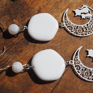 May include: A pair of white earrings with a white oval-shaped stone and a silver crescent moon charm with two cats on it.